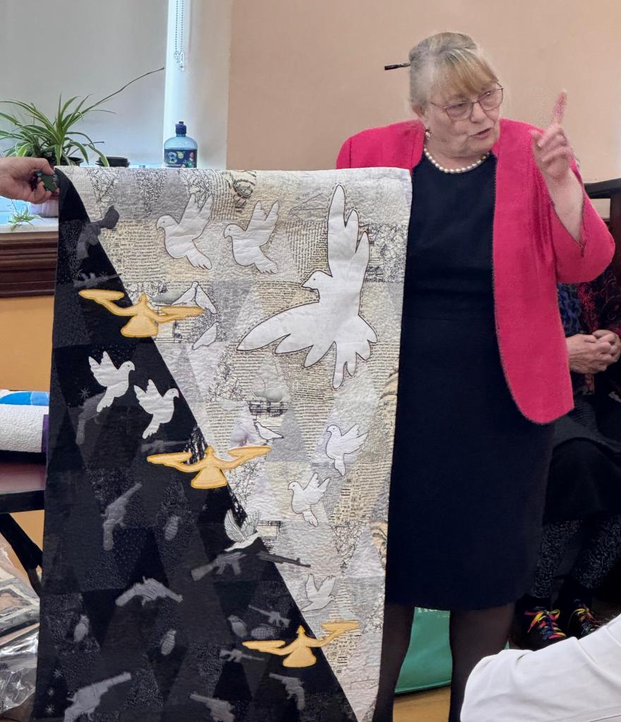 Loretta O'Brien holds her Peace quilt with one side dark, with images of weapons, balanced again a pale side with quilted doves.