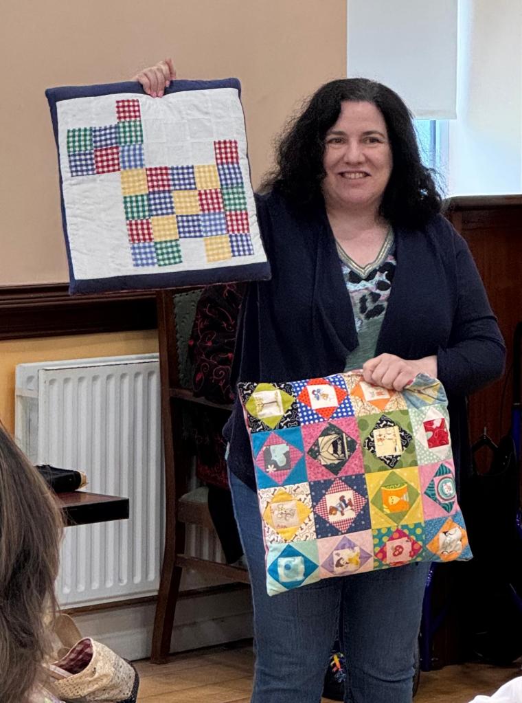 Ruth Bourke holding two cushion covers, one a gingham dog made from squares, the other an economy block swap made of fussy cut centres and surrounding triangles.